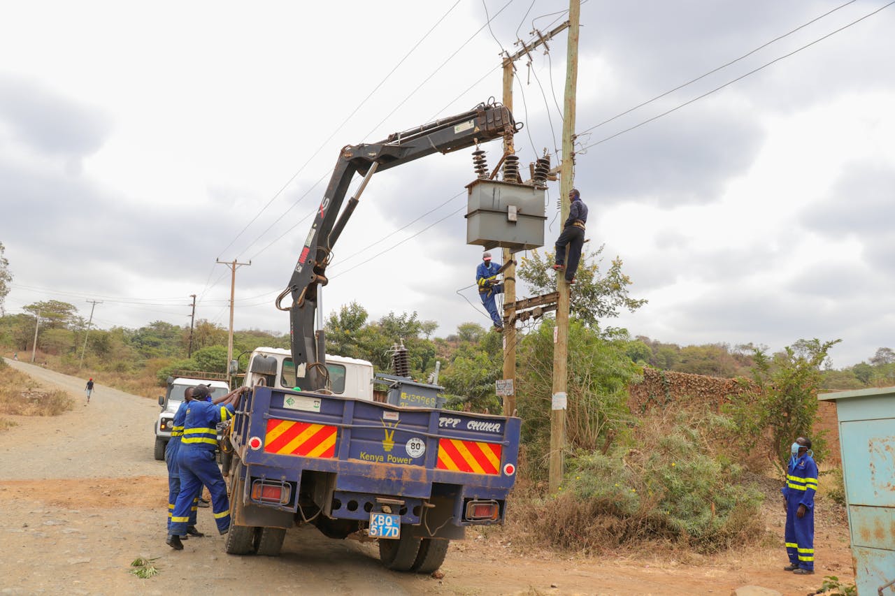 Utility workers in protective gear servicing power lines with a truck crane on a rural road.
